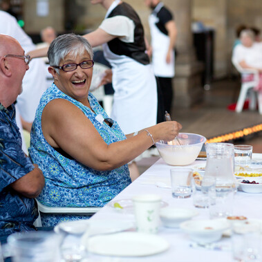 Two audinece members are captured laughing with one another, whilst seated at a dinner table. The women on the right hand side is whisking a bowl of liquid, whilst the man beside her looks on. The table is cluttered with cups, plates and other various bowls.