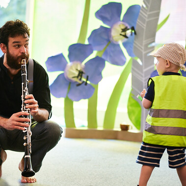 A performer is crouched and playing the clarinet to an inquisitive child in a hi-vis vest.