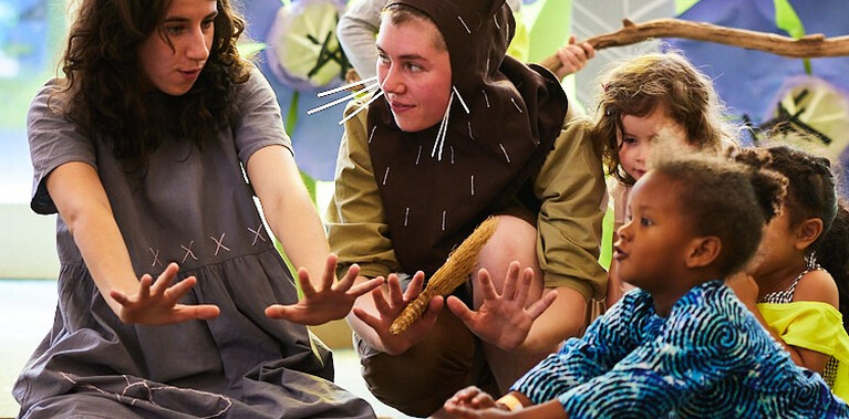 Two performers and a group of children are sat around a pile of sticks. They all have their arms outstretched as if feeling the warmth of an imaginary fire from the pile of sticks.