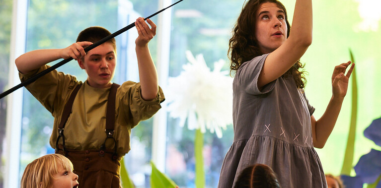 Two performers are photographed surrounded by a group a small kids wearing hi vis vests. The performer on the left is holding a long stick, aimed at head-height. At the end of the stick is a swallow, which the second performer is holding above their head, as if guiding its flight.