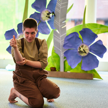 A performer is photographed giving a thumbs-up; behind them are large paper flowers and grass.