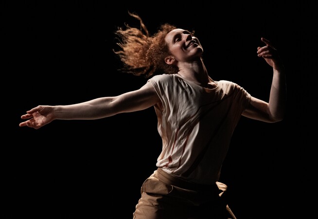 The performer, Nikki Rummer, is photographed mid-dance. Her arms are spread out and her legs are planted wide apart. She is smiling and appears to be liberated through the dance.