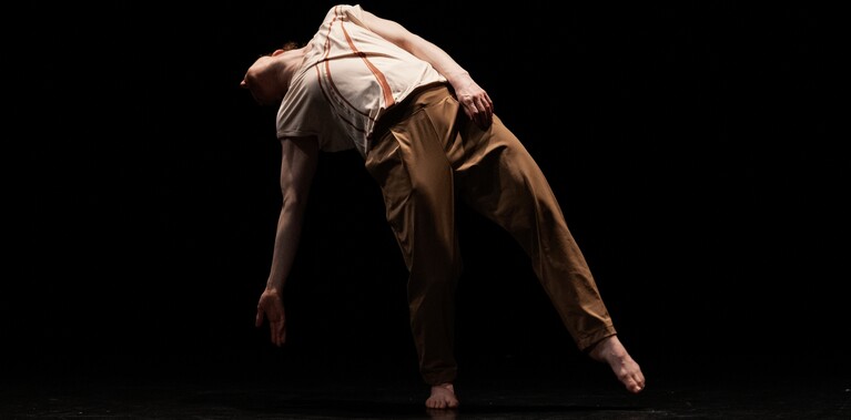 The performer, Nikki Rummer, is photographed in a slumped position, as if her body has given up. She stands on one leg and her right arm reaches towards the ground, with the rest of her body following.