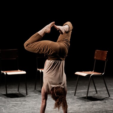 The performer, Nikki Rummer, is photographed mid acrobatic pose. They are stood on their hands, with their legs bent in the air. Behind them are three empty chairs.