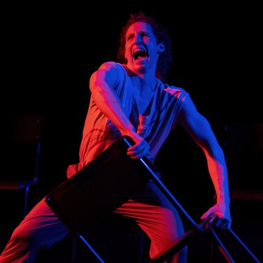 The performer, Nikki Rummer, is photographed grabbing a chair as if they are about to throw it in anger. She is lit by green and blue stage lighting creating a dramatic image.