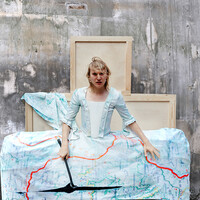 Anders Duckworth stands in a costume that resembles a 15th century dress with a greatly exaggerated skirt made out of a map.