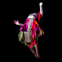 Person wearing a skirt with ribbons in pinks and oranges and leopard print hanging off. They are leaning backwards so their head is not visible. The background is black.