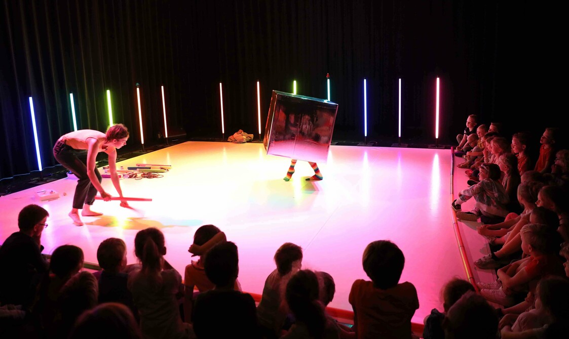 A white stage lit with multi - coloured light, surrounded by glowing multi - coloured sticks. Two figures are the stage; one is crouching down, the other is inside a silver, mirrorlike box with only their feet poking through. A young audience sits around
