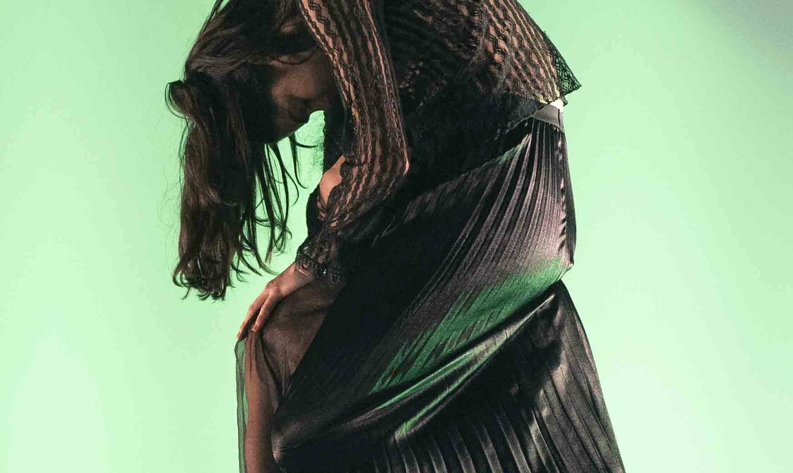 A woman wearing black lace is sitting slumped over the shoulder of someone standing - up, wearing a black see - through shirt. The plain background is lit green.