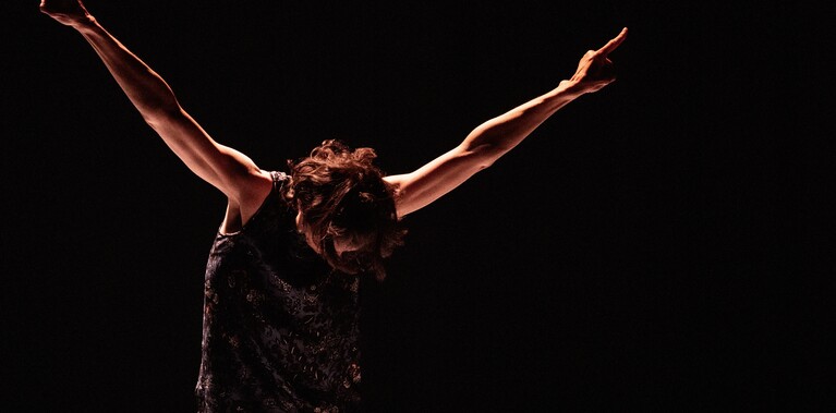 A woman stands in a rock star posture, feet together, creating a figure of a V shape with her arms, her fingers strongly pointing upwards and her head bowed down.