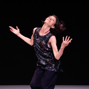 A woman slightly arches her back in an opening gesture of joy whilst her hair floats behind her. She is about 60 and wears a fine silk flowery top and dark navy trousers.