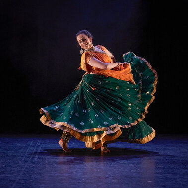 A female Kathak dancer swirls her skirts with her hands. She smiles towards her extended eg. She is wearing a dark green skirt with gold details, with an orange and gold top and pinned dupatta/scarf. She has gold Indian jewellery and white flowers in her hair. The stage is hung with black and the light tints the floor blue. Four female Kathak dancers gesture in unison from a seated tableau position with their right arms extended across their bodies. In the background, two male dancers make a mirrored pose, with opposite arms extended towards each other, and opposite legs extended away. All the dancers wear bright saffron orange and sea green costumes trimmed with gold. The women have gold jewellery and white flowers in their hair. The stage is hung with black and the light tints the floor blue.