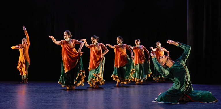Seven Kathak dancers: two male, five female, in bright saffron orange and sea green costumes, performing. The five female dancers are processing diagonally across the stage, holding their skirts out with one hand and gesturing forwards with the other. In the back, a male dancer with orange stands, reaching upwards with one arm and his gaze. In the front a male dancer in green leans backwards from a seated position with both arms raised. The stage is hung with black and the light tints the floor blue.