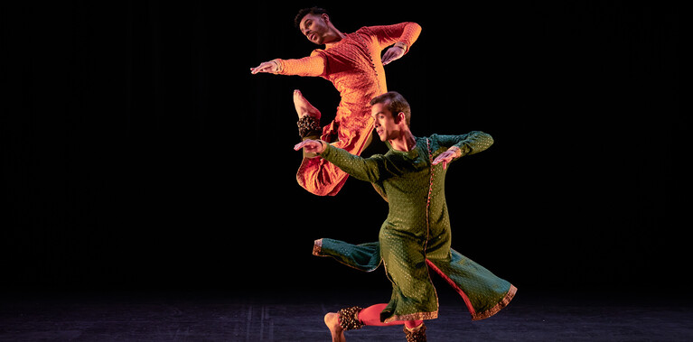 Two male Kathak dancers performing. One is in a saffron kurta, the other in a dark sea green kurta, and both have bells on their ankles. The dancer in orange leaps with legs tucked and arms extended towards a downstage corner. The dancer in green comes out of a spin and extends his arms in the same manner. They form an arrangement of angles that gives the impression of synchronicity. The stage is hung with black and the light tints the floor blue.