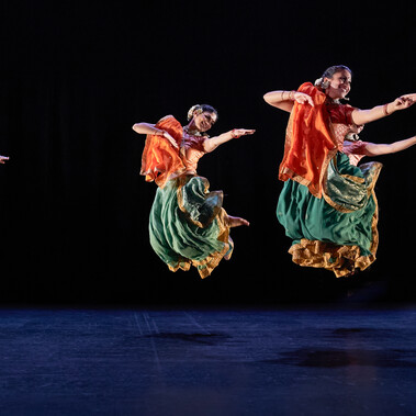 Seven Kathak dancers: two male, five female, in bright saffron orange and sea green costumes, caught mid-leap. They are grouped three towards the back, and four towards the front, but are all dancing in unison. Their legs are tucked up under them, and their arms are all extended in the same direction. Those whose faces are visible are smiling. The women have gold jewellery and white flowers in their hair. The stage is hung with black and the light tints the floor blue.