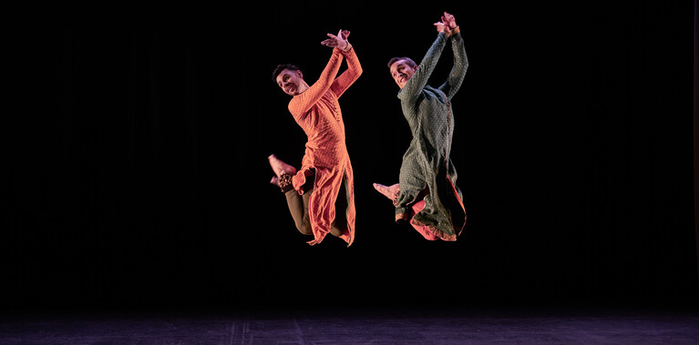 Two male Kathak dancers performing. One is in a saffron kurta, the other in a dark sea green kurta, and both have bells on their ankles. They have been caught mid-leap, with their raised arms crossed at the wrists and their bent legs crossed at the ankles behind them. They are looking over their left shoulders and smiling. The stage is hung with black and the light tints the floor purple.