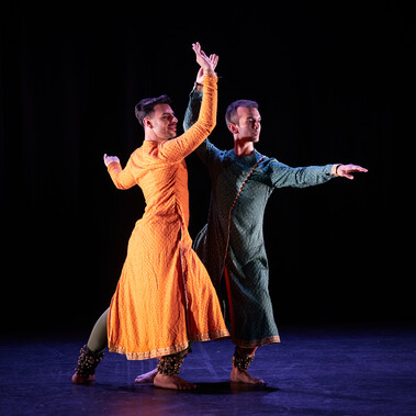 Two male Kathak dancers performing. One is in a saffron kurta, the other in a dark sea green kurta, and both have bells on their ankles. They are standing together lunging to a downstage corner with opposing arms raised and extended. They are looking in the same direction and smiling slightly. The stage is hung with black and the light tints the floor blue.