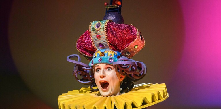 A performer wearing a large colourful crown and yellow collar is pointing to the audience.  They are coming out of a large white ball. The lighting on the backdrop is bright pink.