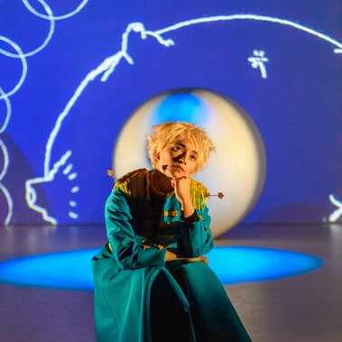 A performer dressed as The Little Prince sits on a stage, looking to the sky. Behind them is a large white ball and white graphic illustrations from the book projected on a blue backdrop.