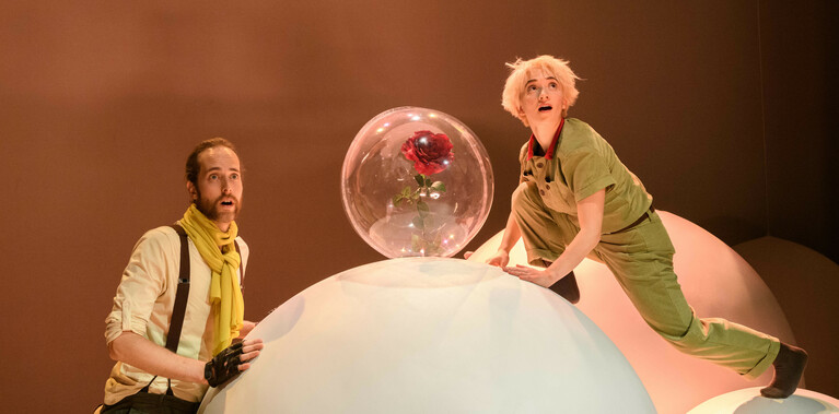 A performer dressed as The Little Prince stands on top of two white balls. One has a glass sphere with a rose in it on top. A performer dressed as The Pilot also stands next to the ball.
