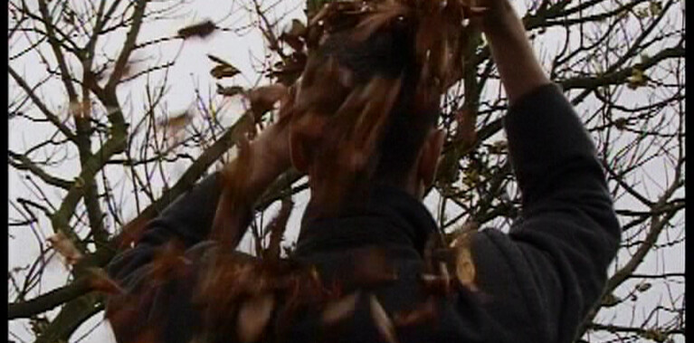 A man in a dark grey overcoat stands with his back to us in a wintery leafless wood, his hands are up over is head amongst the dry crisp autumnal leaves that are falling from his head and flying in the wind