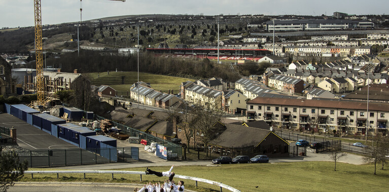 a dancer is tossed in the air by a group of people all looking up at her, arms stretched up to catch her. They are on a Derry street with the panoramic hillside streets of bogside behind them.