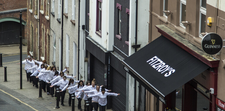 A line of white shirted dancers, arms outstretched as if walking a tightrope, walking up a city street in Derry, just passing a bar that sells Guinness.