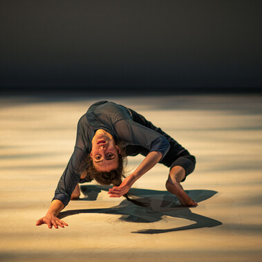 A person performs an expressive dance move, bending backward with one hand on the floor and the other reaching out. The scene is lit dramatically, casting shadows on the light-colored floor.