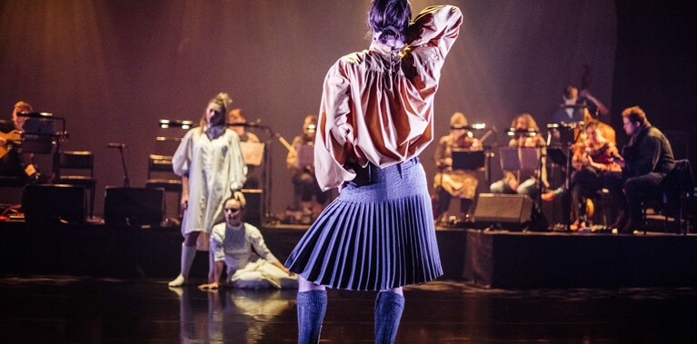 A man facing away from the camera wearing a kilt standing on stage, with one hand in his hair and the other on his hip. Two women, one sitting one standing dressed in white are in the background with orchestra in behind them.