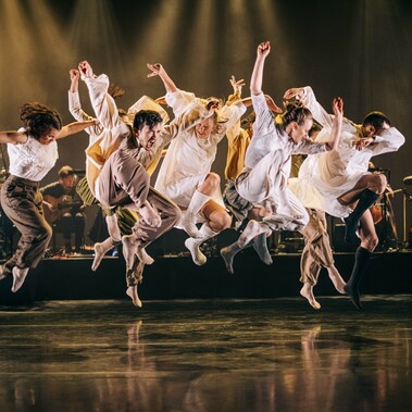 Dancers in white dresses gracefully leaping in the air, showcasing their elegance and skill. In the back ground there is a live band with dark warm lighting.