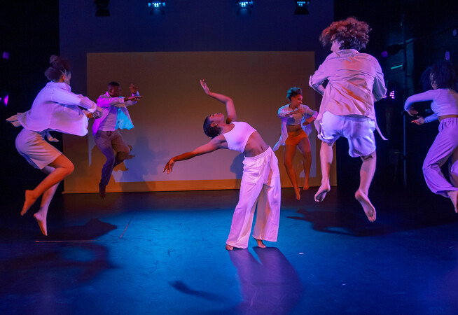 A dancer at the centre of the stage leans back, her arms held gracefully in the air above her head. Around her five dancers leap into the air in a circle.