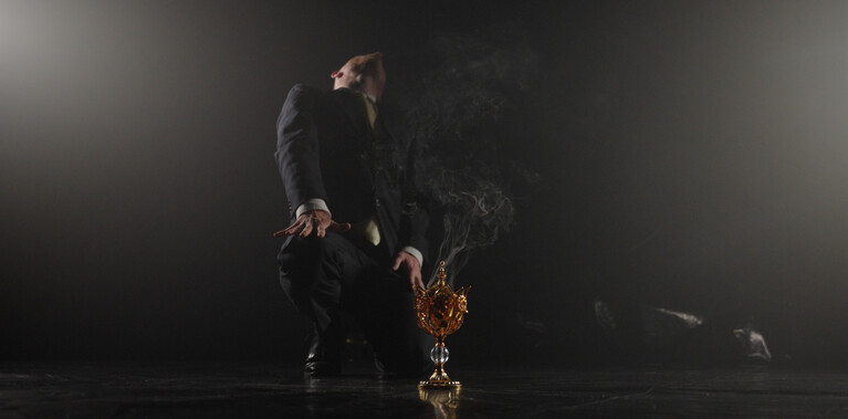 A man dressed in a black suit kneels on a black dance floor in front of a fold vessel, his head is thrown back. There are faint wisps of burning incense coming from the vessel.