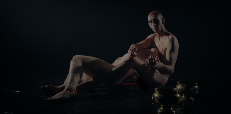 Two nearly identical looking naked men lie together on a black dance floor. Two golden vessels are on the floor in the foreground.