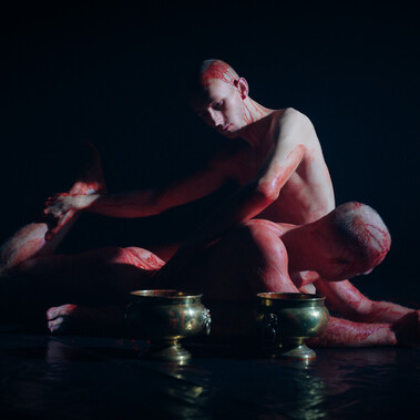 Two nearly identical looking naked men lie together on a black dance floor covered in blood. Two golden vessels are on the floor in the foreground.