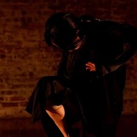 In the image there is a brick wall in the background. A dancer with black hair crouches over their body, lifting their right leg slightly so the foot is by the knee. The dancer is wearing all black.