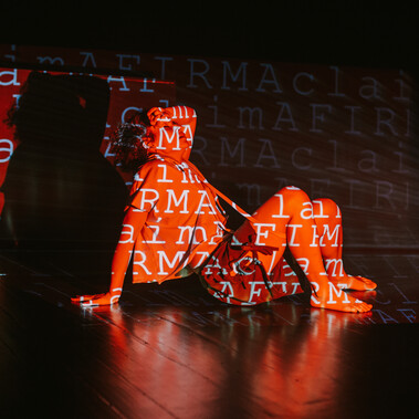 Gaya is sitting in the center of the picture. She is wearing shorts and a sports T-shirt with a hood. Her legs are bent and her feet are flat on the floor. Her torso is slightly tilted backwards, her right hand is resting on the floor and her forearm is resting on her forehead. Her eyes are closed because a text with a red background and white letters is being projected onto her body - and onto the wall behind her - with the word CLAIM repeated several times and in two different languages (English and Portuguese). Above the projection on the wall we see the shadow of Gaya's body. In her expressions we can sense discomfort, tiredness and pain. The intense red of the projection accentuates these sensations.