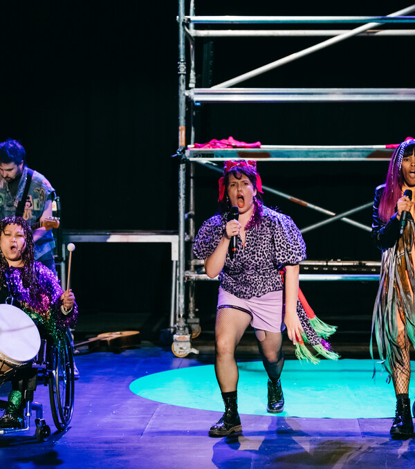 3 band members stand at the front of the stage singing. They are dressed like a punk band, some singing, 1 playing a drum.