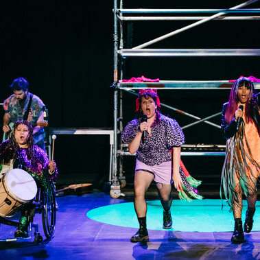 3 band members stand at the front of the stage singing. They are dressed like a punk band, some singing, 1 playing a drum.