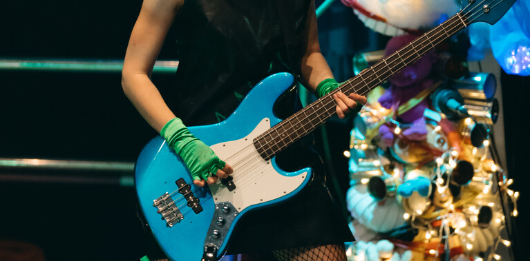 Rake is on stage wearing a punk outfit and headphones, they are playing a bass guitar.