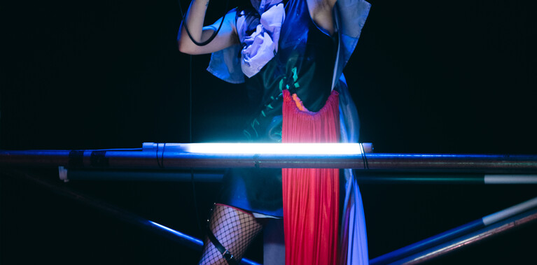 A performer stands on top of a two tier scaff tower singing into a microphone holding 1 arm up. They are wearing a punky costume and headphones.