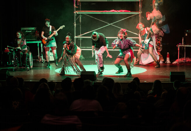 On stage, the band all dressed in punk clothing are rocking out. The full stage which has the backdrop of a scaff tower which red and green light reflects off. The 6 performers are singing, playing bass and guitar and on the drums.
