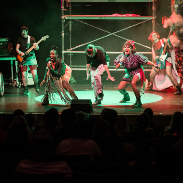 On stage, the band all dressed in punk clothing are rocking out. The full stage which has the backdrop of a scaff tower which red and green light reflects off. The 6 performers are singing, playing bass and guitar and on the drums.