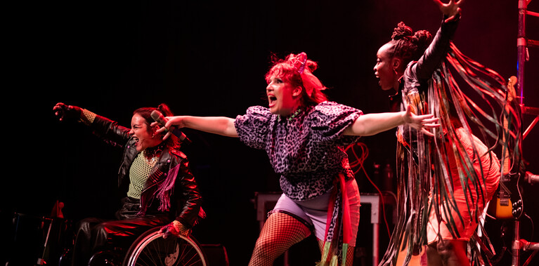 3 performers on stage, dressed in punky costumes of leather, tassels and patterns have their arms and mouths wide as they move energetically across the stage in different directions.