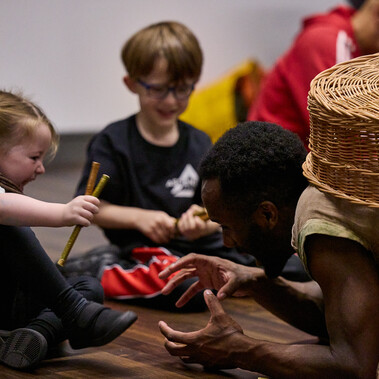 A man is kneeling on the floor next to two children with a woven basket on his back. He reaches for one of the children as she squeals with laughter.