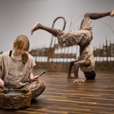 A woman sits on the floor playing a musical instrument in a woven basket. Behind her a man is balancing on his head with his legs in the air.