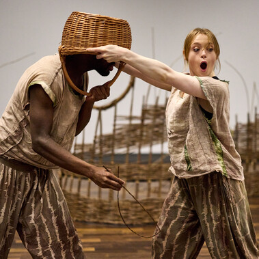 A woman is holding a woven basket over the head of a man holding a fish made from willow. She looks shocked and surprised.