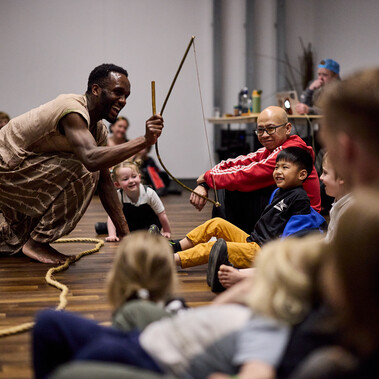 A man is kneeling in front of an audience holding a bow and arrow. They all look excited and happy.