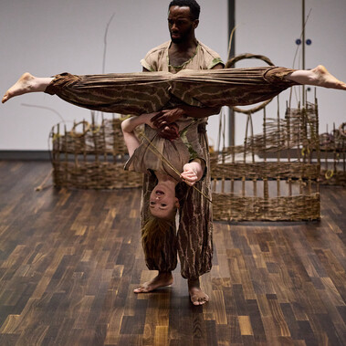 A man holds a woman upside down with her legs spread wide apart. She looks happy and is holding a fish made from willow.