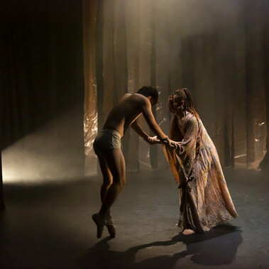 A tall male presenting dancer, dressed only in white shorts is standing on pointe, leaning slightly forward, head bent with his arms outstretched gives the impression of being weak and fragile. He’s leaning in towards a female presenting dancer who is holding both his hands in support and looking intently into his face.