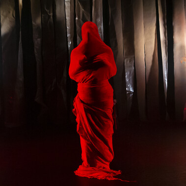 A solo figure wrapped mummy-like from head to toe in bright red coloured cloth stands alone on stage. Both hands are held closely to the chest, the head pointing slightly up and the overall posture conveying the anguish and pleading of someone who has been crushed emotionally.